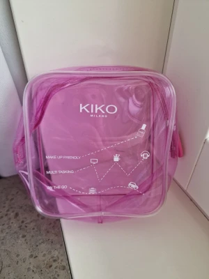Rosa sminkväska från KIKO Milano - Säljer en genomskinlig rosa sminkväska från KIKO Milano. Väskan är fyrkantig med dragkedja och har tryckt text och ikoner på framsidan. Perfekt för att förvara sminkborstar, läppstift eller andra sminkprodukter. Materialet är plast och väskan är lätt att rengöra. Använd en eller två gånger, jättefint skick