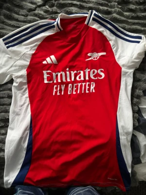 Arsenal matchtröja Adidas röd/vit - Säljer en Arsenal matchtröja från Adidas med klassisk röd front, vita ärmar och blå detaljer. Tröjan har klubbmärke och Adidas-logga på bröstet samt 'Emirates Fly Better' tryckt framtill. Tillverkad i lätt och ventilerande polyester, perfekt för fotboll.