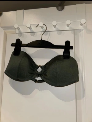 Mörkgrön bandeau bikiniöverdel H&M - Snygg mörkgrön bandeau bikiniöverdel från H&M med en rund detalj i mitten och justerbart spänne bak. Stilren och enkel design i mjukt syntetmaterial, perfekt för strand eller pool.