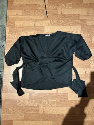 Svart omlottopp med knytband - Stilren svart topp i omlottmodell med korta ärmar och knytband i sidan som ger en smickrande passform. Tillverkad i mjukt och följsamt tyg, bekväm att bära både till vardags och vid festligare tillfällen. Kan enkelt kombineras med jeans, kjol eller byxor.