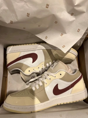 Nike Air Jordan 1 Low beige/vinröd - Nike Air Jordan 1 Low sneakers i beige, vitt och vinrött. Skorna har en klassisk låg siluett med vinröd swoosh, snörning och detaljer i både slätt och texturerat skinn. Perfekta för dig som vill ha en clean men ändå unik look.