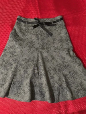 Grå blommig kjol från Orsay XS  - Supersnygg grå kjol från Orsay med blommigt och rutigt mönster. Kjolen har en svart rosett i midjan och lätt utsvängd form. Perfekt för dig som gillar unika detaljer och vill sticka ut lite extra.