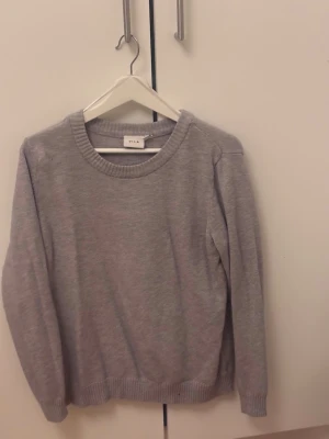 Grå stickad tröja från VILA - En enkel och stilren grå stickad tröja från VILA. Tröjan har rund halsringning, ribbade muddar vid ärmslut och nederkant samt lång ärm. Perfekt för lager på lager under höst och vinter.