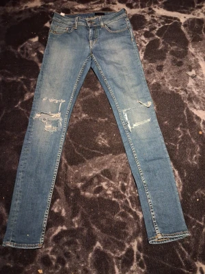 Ljusblå jeans från Tiger of Sweden med slitningar  - Säljer ett par ljusblå skinny jeans från Tiger of Sweden, modell Janet. Jeansen har slitna detaljer och hål på båda benen för en cool, avslappnad look. Klassisk femficksmodell med gylf och knapp, tillverkade i Italien. Perfekta till sneakers eller boots.