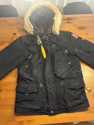 Svart Parajumpers parkas med päls - Säljer en svart Parajumpers parkas med huva och päls, ikonisk gul rem vid halsen och flera praktiska fickor. Jackan har dragkedja och knappar framtill, patch på ärmen och quiltad insida med orange detaljer. Perfekt för kalla dagar.