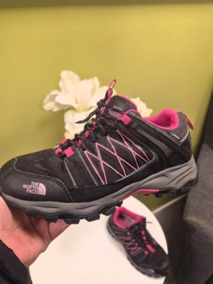 Svarta sneakers från The North Face 39 - Säljer ett par svarta sneakers från The North Face med rosa detaljer och grå sula. Skorna har snörning, meshpaneler för ventilation och slitstark syntet på ovandelen. Perfekta för dig som gillar outdoor och vill ha en sportig look.