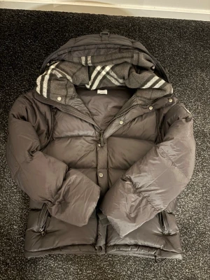 Svart pufferjacka från Burberry - Säljer en svart pufferjacka från Burberry med avtagbara ärmar så den kan användas som väst. Jackan har huva med grått rutigt foder, dragkedja och knappar framtill samt två fickor med dragkedja. Klassisk Burberry-logga på ena ärmen.