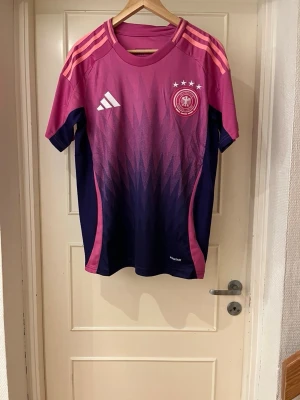 Germany away shirt 24/25 - Säljer en Adidas Germany away matchtröja i storlek S. Tröjan har en snygg rosa och mörkblå färgskala med grafiskt mönster, tre ränder på axlarna och broderad landslagsemblem. Tillverkad i polyester och perfekt för fotboll eller träning.