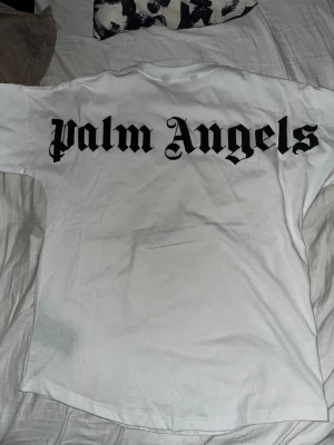 Vit Palm Angels t-shirt med tryck - Säljer en clean vit t-shirt från Palm Angels med stort svart logotyp-tryck på ryggen och mindre tryck vid halsen. Klassisk rund hals och korta ärmar. Skön och mjuk bomull, perfekt till streetwear-looken. Pris kan diskuteras den är oanvänd.