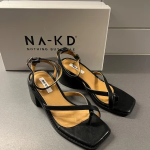 Svarta sandaler med klack från NA-KD - Snygga svarta sandaler från NA-KD med bred klack och fyrkantig tå. Skorna har tunna remmar i skinnimitation som korsas över foten och justerbart spänne vid ankeln. Perfekta till sommarens outfits.