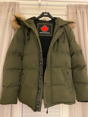 Grön pufferjacka från Zavetti Canada - Säljer en grön pufferjacka från Zavetti Canada med stor huva och fuskpälsdetalj. Jackan har dragkedja, tryckknappar och stora fickor framtill. Insidan är quiltad och fodrad för extra värme. Perfekt för kalla dagar och riktigt snygg streetstil.