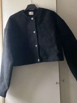 Svart jacka från H&M - Säljer en svart jacka från H&M med croppad passform. Jackan har rund halsringning, långa ärmar och stängs med silvriga knappar framtill. Perfekt för dig som gillar en enkel men trendig look.