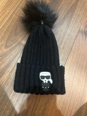 Svart stickad mössa med tofs - Svart ribbstickad mössa med stor fluffig fuskpäls-tofs på toppen och en cool patch framtill med en tecknad figur i kostym. Perfekt accessoar för att hålla värmen och addera lite edge till din outfit.