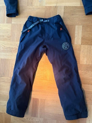 Svarta täckbyxor från Dope Snow - Säljer ett par svarta täckbyxor från Dope Snow med elastisk midja och justerbart bälte. Knappt använda o i väldigt bra skick. Säljer även en jacka som här till