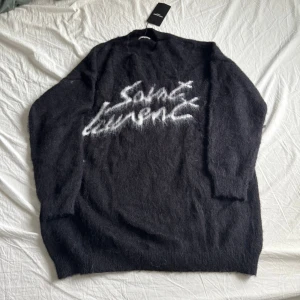 Svart Saint Laurent stickad tröja - Svart stickad tröja från Saint Laurent med vit graffiti-inspirerad logga framtill. Tröjan har lång ärm, rund hals och en lite oversized passform. Mjuk och fluffig känsla, perfekt för kyliga dagar. Funkar som XL