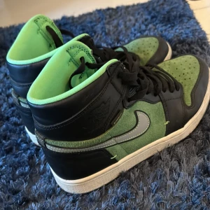 Nike Air Jordan 1 High svart/grön - Nike Air Jordan 1 High sneakers i svart och grön mocka med svarta läderdetaljer och svart snörning. Swoosh-logga i silver på sidan och vit sula med genomskinlig undersida. Klassisk high-top siluett som sticker ut med sin färgkombination.