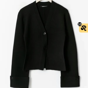 Svart kofta från Gina Tricot - Stilren svart kofta från Gina Tricot med v-ringad halsringning och två knappar framtill. Långärmad modell med breda muddar och knappdetalj vid ärmslut. Perfekt för lager-på-lager och enkel att matcha med allt. Använd en gång