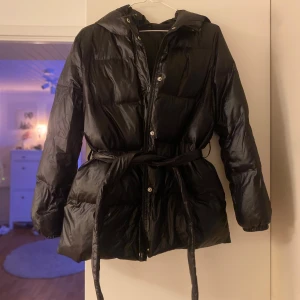 Svart pufferjacka med bälte från SHEIN - Svart pufferjacka från SHEIN i storlek 38. Jackan har huva, bälte i midjan och dragkedja framtill. Den är quiltad och har en glansig finish som ger en cool vibe. Perfekt för kalla dagar när du vill vara både varm och snygg.