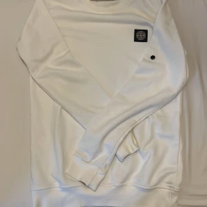Vit långärmad tröja Stone Island. Den har ingen stone Island märke på armen - Snygg vit långärmad tröja från Stone Island med klassisk rund hals och ikonisk logopatch på bröstet. Tröjan har en clean look och är gjord i mjuk bomull, perfekt för dig som gillar stilrena plagg med streetkänsla. 