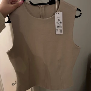 Beige ärmlös topp från Gina Tricot - Snygg beige topp från Gina Tricot med rund halsringning och ärmlös design. Toppen har en stilren look med diskreta sömdetaljer framtill och dragkedja i ryggen. Perfekt att styla med jeans eller kjol för en clean vibe.