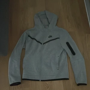 Grå Nike vindjacka med huva - Snygg grå vindjacka från Nike med svart logga på bröstet och svarta detaljer. Jackan har hel dragkedja, huva och en cool ficka med dragkedja på ena ärmen. Perfekt för dig som gillar sportig stil och vill ha något bekvämt och stilrent.