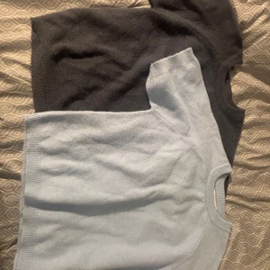 Två stickade kortärmade tröjor - Säljer två stickade tröjor med kort ärm, en i ljusblått och en i mörkgrått. Båda har rund halsringning och ribbade kanter. Perfekta för lager-på-lager och chill stil.