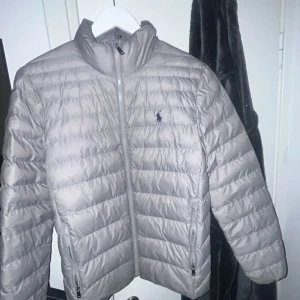 Ljusgrå dunjacka från Ralph Lauren - Snygg ljusgrå dunjacka från Ralph Lauren med klassisk quiltad design och dragkedja framtill. Jackan har hög krage, två fickor med dragkedja och den ikoniska loggan broderad på bröstet. Perfekt för kyliga dagar.