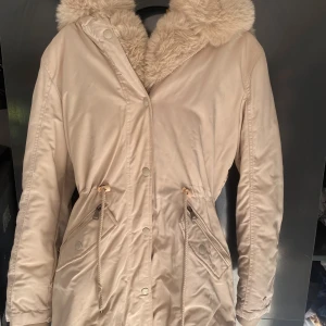 Beige parkasjacka med fuskpäls - Snygg beige parkasjacka med stor fluffig fuskpäls runt huvan. Jackan har dragsko i midjan, två stora fickor med dragkedja och knappar samt långa ärmar. Perfekt för kyliga dagar och ger en riktigt mysig vibe.