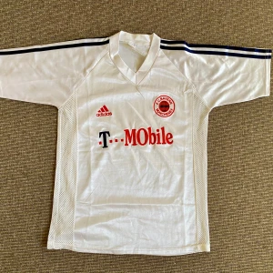 Vit FC Bayern München Adidas tröja - Säljer en klassisk vit FC Bayern München fotbollströja från Adidas med T-Mobile sponsortryck. Tröjan har korta ärmar, svarta ränder på axlarna och meshpaneler på sidorna för extra ventilation. Broderad klubbmärke och Adidas-logga framtill. Tillverkad i polyester. Skador finns, se bilder