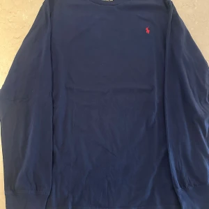 Mörkblå långärmad tröja Polo Ralph Lauren - Mörkblå långärmad tröja från Polo Ralph Lauren i bomull. Klassisk design med rund hals och röd broderad logga på bröstet. Perfekt för en stilren och avslappnad look. Passar dig som gillar enkel men snygg stil. Tröjan på bild nr2 är lite ludd på men allt det äe borttaget nu i efterhand.