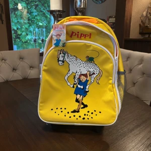 Gul Pippi ryggsäck med hjul - Säljer en färgglad gul ryggsäck med motiv av Pippi Långstrump, hennes häst och apa. Väskan har praktiska hjul och utdragbart handtag, perfekt för att rulla med sig. Framsidan har ett stort fack med dragkedja och meshficka på sidan. Superfin för dig som gillar Pippi! Helt ny med prislapp kvar 