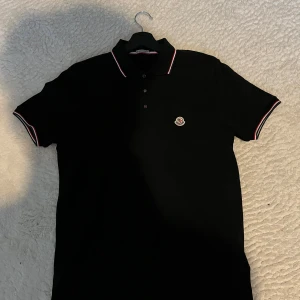 Svart Moncler pikétröja med röd/vit kant - Svart pikétröja från Moncler med klassisk krage och knäppning framtill. Tröjan har röda och vita ränder på krage och ärmslut samt Moncler-logga broderad på bröstet. Tillverkad i bomull för en skön och stilren känsla. Priset är inte hugget i sten vid snabb affär, men priset är bra jämfört med ny från affär.