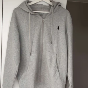 Grå hoodie från Ralph Lauren - Snygg grå hoodie från Ralph Lauren med dragkedja framtill, huva med snörning och klassisk logga på bröstet. Tröjan har känguruficka och är tillverkad i mjuk bomull. Perfekt för en chill och stilren look.