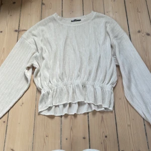 Ribbad beige topp med volang från Zara - Superfin ribbad topp från Zara i beige med lång ärm och volangdetalj nedtill. Modellen är croppad med resår i midjan för en snygg passform. Perfekt till jeans eller kjol för en trendig look.