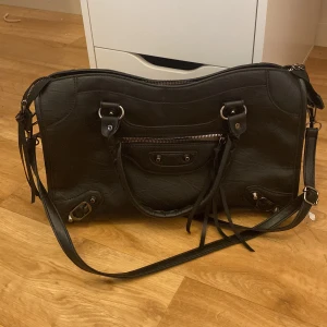 Svart axelväska i skinnstil med dragkedja - Cool svart axelväska i skinnimitation med både handtag och lång axelrem. Väskan har snygga metalldetaljer, dragkedja upptill och framtill samt dekorativa remmar. Insidan är ljus och rymlig, perfekt för att få plats med allt du behöver.