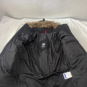 Svart parkas från Canada Goose L - Svart parkas från Canada Goose med äkta päls på huvan och klassisk logga på ärmen. Jackan är lång, har dragkedja och knappar framtill samt flera innerfickor. Perfekt för kalla dagar med sin dunfyllda insida och snygga detaljer.