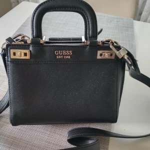 Svart handväska från Guess med gulddetaljer - Snygg svart handväska från Guess med guldfärgade metalldetaljer och dragkedja. Väskan har både handtag och axelrem, samt flera fack inuti för smart organisering. Tillverkad i skinnimitation och har stilren, boxig form. Perfekt för dig som vill ha en trendig och praktisk väska. Tyget har lossnat lite   (ca 1 cm)'från sömnen inuti 