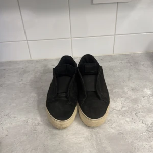 Svarta sneakers från Axel Arigato (använda) - Svarta sneakers från Axel Arigato i mocka med chunky vit sula. Slip-on modell utan snörning för en clean look. Snygg minimalistisk design med rund tå och diskret logga på sidan. Perfekta för dig som gillar stilrena och trendiga skor.