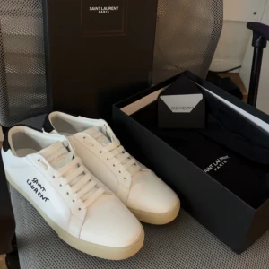 Saint Laurent vita sneakers - Snygga vita sneakers från Saint Laurent med svart detalj på hälen och handskriven logga på sidan. Skorna är i slätt skinn med vit snörning och krämfärgad sula. Perfekta för dig som gillar stilrena och exklusiva sneakers med en touch av lyx. Använd fåtal gånger, går att förhandla.