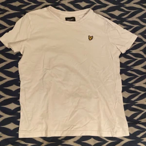 Vit t-shirt från Lyle & Scott Junior (9-10 år) - Vit t-shirt från Lyle & Scott Junior med klassisk rund hals och korta ärmar. På bröstet sitter den ikoniska broderade gula örnen. Skön och enkel modell i mjuk bomull, perfekt till jeans eller shorts. I storlek 9-10 år.