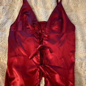 Röd satin topp med snörning - Snygg röd topp i glansig satin med smala axelband och djup v-ringning. Framsidan har dekorativa snörningar som ger en unik look. Perfekt för dig som vill sticka ut med en elegant och trendig stil.