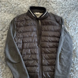Grå cardigan jacka från Moncler XL - Sitter som L! Snygg grå cardigan jacka från Moncler i storlek XL. Jackan har quiltad front i mörkgrått med dragkedja, ärmar och rygg i mjuk bomull, samt ribbad krage med vita ränder. Perfekt för dig som vill ha en sportig och stilren look.