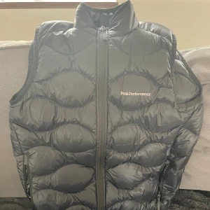 Svart dunväst från Peak Performance - Snygg svart dunväst från Peak Performance med vågformat quiltat mönster och hög krage. Västen har dragkedja framtill och två sidofickor med dragkedja. Perfekt för lager-på-lager och ger en sportig vibe.