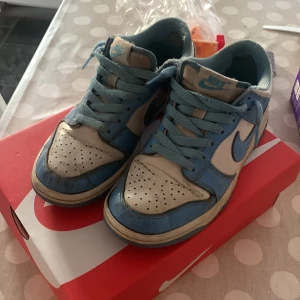 Nike Dunk Low blå/vita sneakers - Nike Dunk Low sneakers i blått och vitt med klassisk låg siluett. Skorna har blå snörning, perforerad tå och vit bas med blå detaljer och swoosh. Tillverkade i skinn och syntetmaterial, perfekt för dig som gillar streetwear och vill sticka ut med en ikonisk modell.