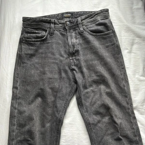 Grå jeans från Jack & Jones, 29/30 - Säljer ett par grå jeans från Jack & Jones i klassisk femficksmodell. Jeansen har en snygg tvättad look, raka ben och normal passform. Tillverkade i slitstarkt denim med bälteshällor och knappgylf. Perfekta för en avslappnad stil.