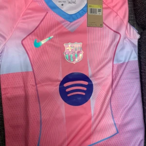 FC Barcelona x Nike rosa fotbollströja S - Snygg FC Barcelona fotbollströja från Nike i rosa och blå toner. Tröjan har korta ärmar, V-ringning med blå kant och tryckta loggor från både Nike och Spotify. Tillverkad i lätt och ventilerande Dri-Fit-material, perfekt för match eller träning.