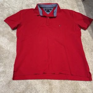 Röd pikétröja Tommy Hilfiger XL - Röd pikétröja från Tommy Hilfiger i storlek XL. Klassisk krage med blå/vit randigt tyg på insidan, liten broderad logga på bröstet och knappar framtill. Tillverkad i bomull för en skön och luftig känsla.