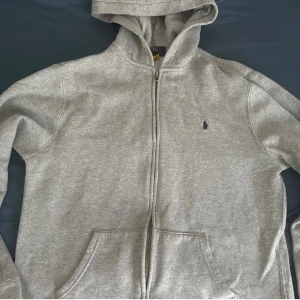 Grå hoodie från Polo Ralph Lauren XL - Klassisk grå hoodie från Polo Ralph Lauren i storlek XL. Tröjan har dragkedja, huva och kängurufickor framtill. Broderad logga på bröstet och mjukt, fleecefodrat innerfoder. Perfekt för chill dagar och enkel att matcha med jeans eller joggers.