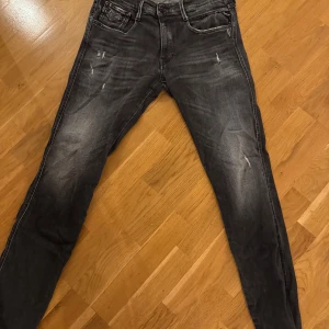 Replay Anbass svarta slim jeans - Svarta Replay Anbass jeans i slim fit med slitningar och snyggt tvättad look. Klassisk femficksmodell med dragkedjegylf och diskreta detaljer. Perfekta för dig som gillar en smalare passform och streetinspirerad stil. Storlek 33/34
