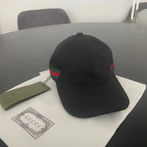 Svart Gucci keps - Svart keps från Gucci i bomull med diskret GG-monogram över hela kepsen. Klassisk böjd skärm och justerbar rem baktill. Sidorna har Guccis ikoniska gröna och röda ränder. Perfekt accessoar för en trendig streetstyle-look.
